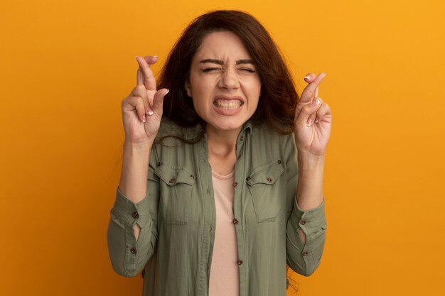 Preocupado por los ojos cerrados joven hermosa vestida con camiseta verde oliva cruzando los dedos aislados en la pared amarilla