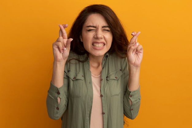 Preocupado por los ojos cerrados joven hermosa vestida con camiseta verde oliva cruzando los dedos aislados en la pared amarilla