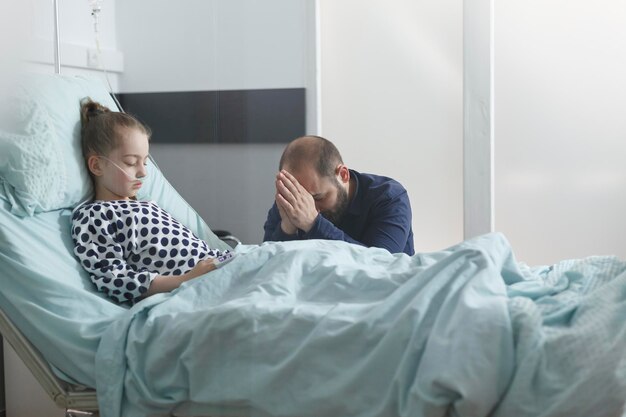 Preocupado padre joven inquieto orando por una paciente enferma hospitalizada mientras está en la sala médica. Niña enferma que se recupera de la enfermedad mientras respira a través del tubo de oxígeno.