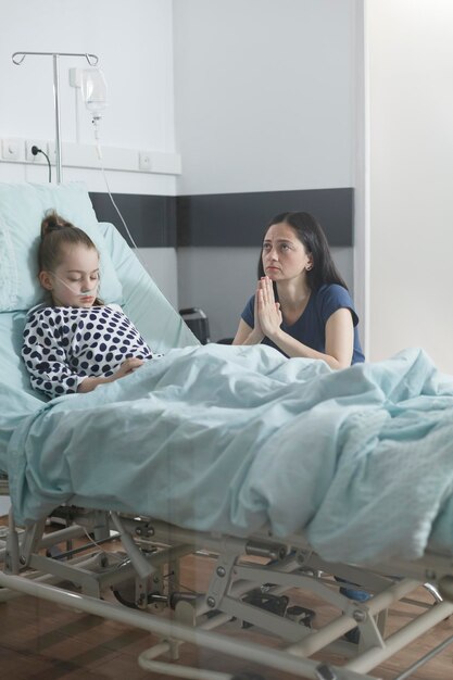 Preocupado padre triste rezando por los enfermos hospitalizados en la sala de la clínica pediátrica. Infeliz mujer atenta rezando por la mala salud de su hija enferma en la habitación del paciente del hospital.