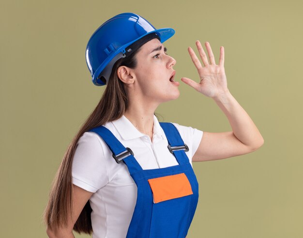 Preocupado de pie en la vista de perfil joven constructor de uniforme llamando a alguien aislado en la pared verde oliva