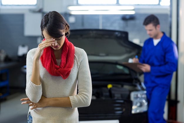 Preocupados cliente permanente, mientras que el examen de coche mecánico en el fondo