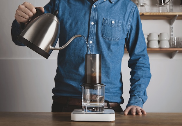 Preparación de café aeropress paso a paso Barista en camisa de jeans vierte agua hervida caliente de la tetera a Aeropress Cafetería profesional de preparación de café