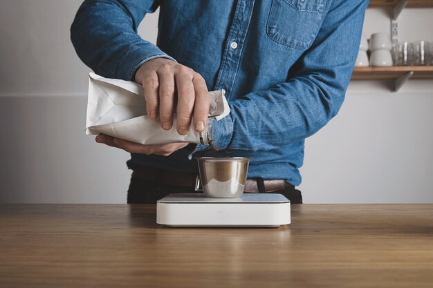 Preparación de café con prensa aerodinámica paso a paso Barista con camisa de jeans vierte granos tostados de la bolsa a la taza de acero sobre pesas blancas Cafetería profesional de elaboración de café
