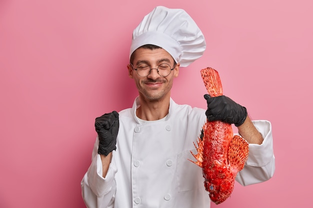 Preparando mariscos. Feliz chef europeo en uniforme de cocinero, guantes de goma sostiene lubina roja, aprieta el puño con alegría