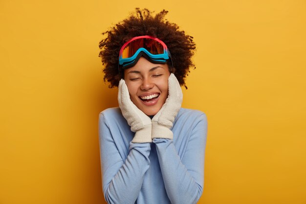 Preparándose para el invierno. Alegre joven rizada en ropa de snowboard, mantiene ambas manos en las mejillas, usa guantes calientes, recuerda un agradable momento de vacaciones