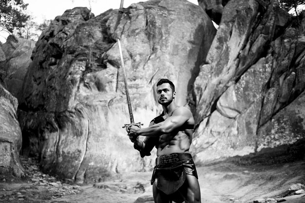 Preparándose para su batalla. Retrato monocromo de un valiente guerrero varonil con una espada posando valientemente cerca de las rocas