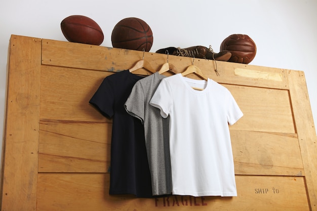 Presentación de una camiseta lisa blanca, gris y negra de manga corta con fútbol vintage, baloncesto y voleibol y botas deportivas viejas de cuero encima de una caja de envío de madera