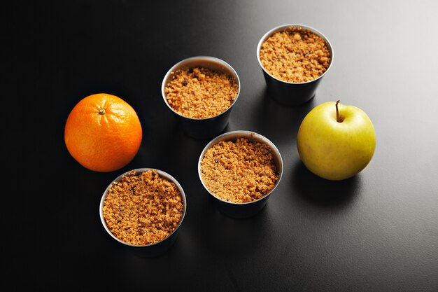 Foto gratuita presentación de cuatro tazas idénticas de acero inoxidable con postre de crumble de manzana, una manzana naranja y una manzana amarilla tomada desde la parte superior sobre una mesa negra
