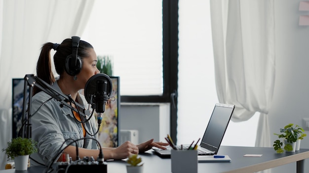 Presentador de programas de radio por Internet hablando por micrófono mientras revisa la lista de temas en una laptop. Popular creador de contenido de medios sociales hablando con la audiencia mientras está sentado en un estudio moderno.