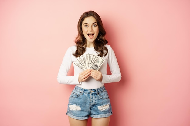 Foto gratuita préstamos y microcréditos. hermosa chica sonriente mostrando dinero, dinero en efectivo en las manos y luciendo entusiasta, de pie sobre un fondo rosado.