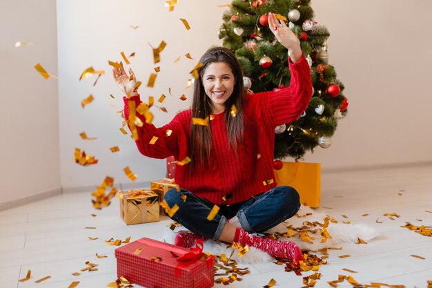 Pretty Woman en suéter rojo sentado en casa en el árbol de Navidad lanzando confeti dorado rodeado de regalos y cajas de regalo