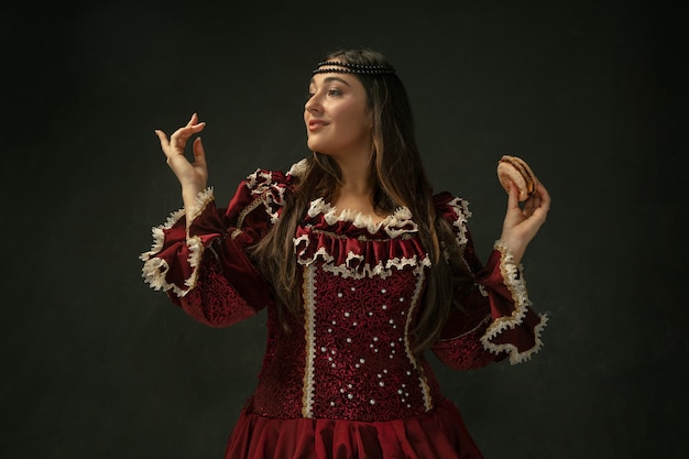 Primer amor. Retrato de mujer joven medieval en ropa vintage roja con hamburguesa sobre fondo oscuro. Modelo femenino como duquesa, persona real. Concepto de comparación de épocas, moderno, moda, belleza.