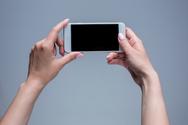 Primer disparo de una mujer escribiendo en el teléfono móvil sobre fondo gris. Manos femeninas sosteniendo un teléfono inteligente moderno y apuntando con el dedo.