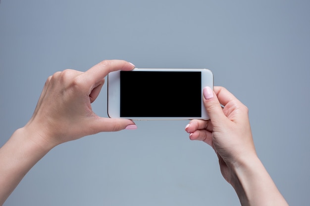 Primer disparo de una mujer escribiendo en el teléfono móvil sobre fondo gris. Manos femeninas sosteniendo un teléfono inteligente moderno y apuntando con figer. Pantalla en blanco para ponerla en su propia página web o mensaje.