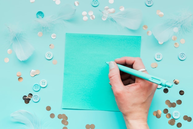 Primer de la escritura de la mano de la mujer en el papel con la pluma rodeada con la pluma; lentejuelas y botón sobre fondo verde azulado