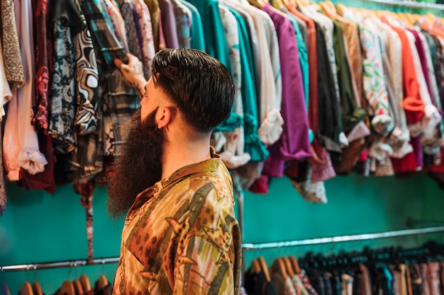 Primer del hombre joven barbudo que mira las camisas que cuelgan en el carril en la tienda