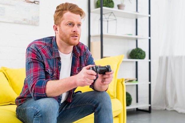 Primer del hombre joven que se sienta en el sofá amarillo que juega al videojuego con la palanca de mando en la sala de estar