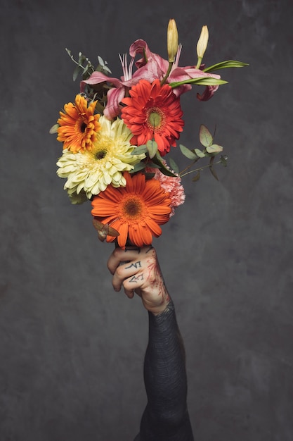 Primer del hombre joven tatuado que sostiene el ramo floral disponible contra la pared gris