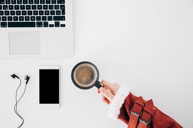 Primer de la mano de la hembra que sostiene la taza de café sobre el escritorio de oficina con el ordenador portátil; telefono movil y auricular