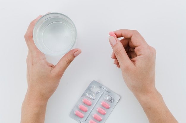 Primer de la mano de la mujer que sostiene píldoras y el vidrio rosados de agua en el fondo blanco