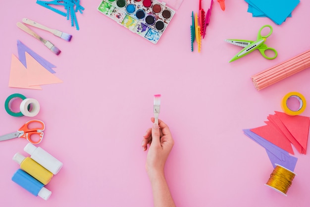 Primer de la mano de la mujer que sostiene el pincel con la paleta de colores del agua; Cepillo de pintura; papel; tijera sobre fondo rosa