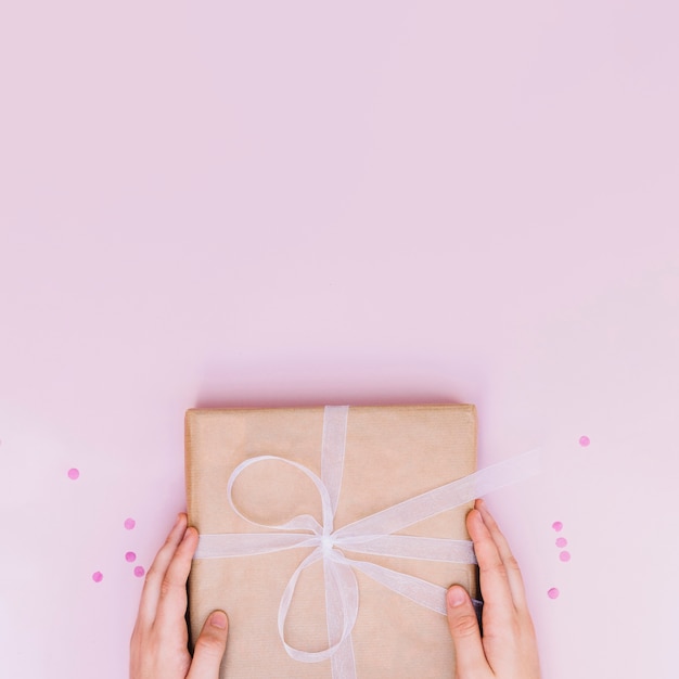 Foto gratuita el primer de la mano que lleva a cabo cumpleaños envolvió la caja de regalo con la cinta blanca en fondo rosado