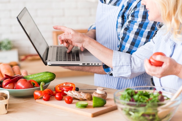 Foto gratuita el primer de la mujer mayor que señala en el ordenador portátil se sostiene por su marido mientras que prepara la ensalada vegetal