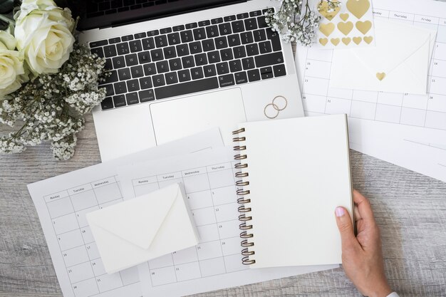 Primer de una persona que sostiene el cuaderno espiral en blanco con la computadora portátil; anillos de boda; flor; Sobre y calendarios en escritorio de madera.