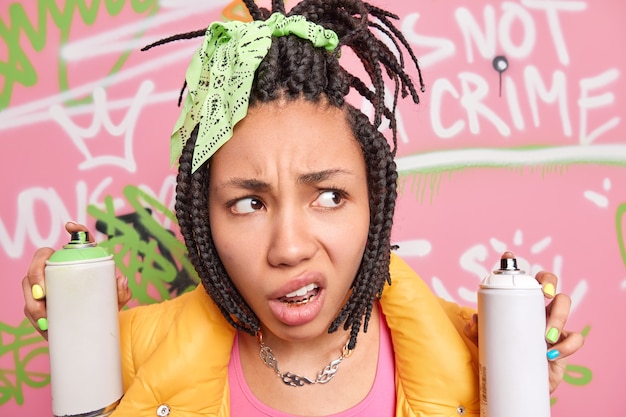 Foto gratuita primer plano de una adolescente afroamericana indignada e insatisfecha con los labios concentrados a un lado tiene peinado dreadlock sostiene botellas de aerosol hace pintura creativa en la pared de la calle