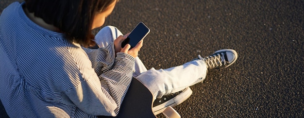 Foto gratuita primer plano de una adolescente patinadora sentada en un monopatín con un mensaje en un teléfono inteligente mirando su teléfono móvil