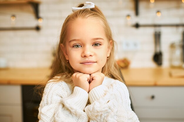 Primer plano de una adorable niña con ojos verdes y cabello largo y suelto tomados de la mano debajo de la barbilla con una sonrisa posando en la cocina