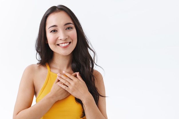 Primer plano agradecido, tierna joven del este de Asia que se siente conmovida y halagada, sonriendo y mirando con afecto, toca el corazón, expresa gratitud y deleite de recibir un bonito regalo