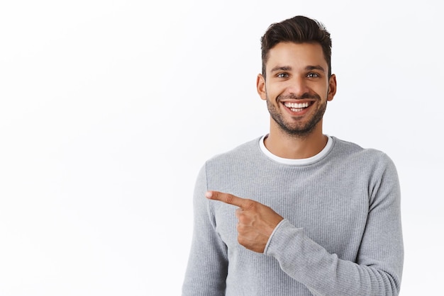 Primer plano alegre apuesto hombre moderno con barba saliente en suéter gris pidiendo café apuntando a la izquierda y sonriendo invitando a ver ver nueva promoción recomendar banner corporativo