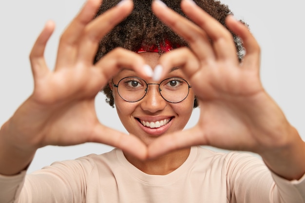 Primer plano de alegre despreocupada encantadora chica de piel oscura hace signo de mano en forma de corazón sobre la cara