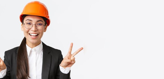 Primer plano de una alegre y exitosa ingeniera asiática arquitecta de la construcción en casco de seguridad y traje de negocios mostrando signos de paz y sonriendo kawaii de pie fondo blanco