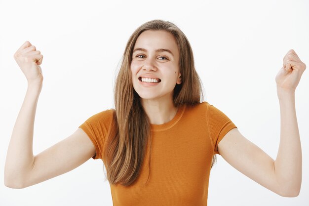 Primer plano de alegre linda joven regocijándose, levantando las manos en gesto de hurra, bomba de puño y sonriendo, triunfando sobre la victoria