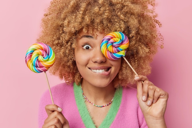 Primer plano de una alegre modelo femenina de pelo rizado que sostiene dos deliciosas piruletas multicolores que muerde los labios se divierte, no le importan los efectos nocivos del azúcar aislado sobre fondo rosa