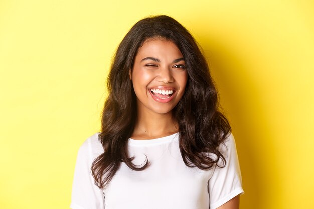 Primer plano de una alegre niña afroamericana, guiñando un ojo y sonriendo a la cámara, recomendando algo bueno, de pie sobre fondo amarillo