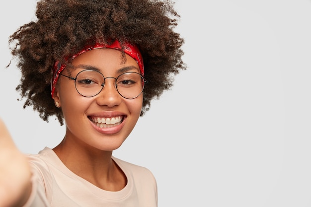 Primer plano alegre niña femenina tiene una sonrisa con dientes, peinado afro, hermosa piel limpia, usa lentes transparentes, estira la mano mientras sostiene un dispositivo irreconocible, hace selfie sobre una pared blanca