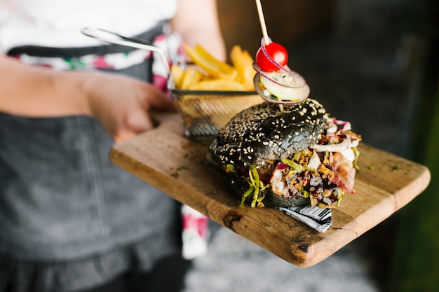 Primer plano de alto ángulo de camarero con tablero de madera con hamburguesa negra y papas fritas