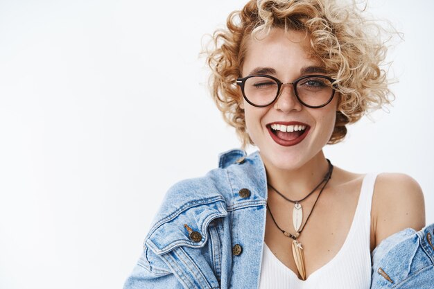 Primer plano de una amiga caucásica moderna con estilo sociable alegre y emocionada despreocupada con corte de pelo corto y rizado en gafas y mezclilla guiñando un ojo y sonriendo coqueta sobre la pared blanca