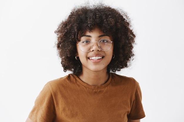 Primer plano de una amigable mujer afroamericana de buen aspecto con gafas transparentes y camiseta marrón sonriendo con una sonrisa alegre y agradable, satisfecha con cómo van las cosas sobre la pared gris