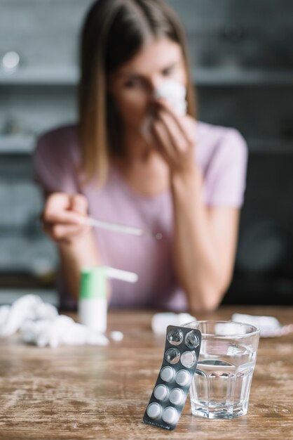 Primer plano de ampolla con pastillas y vaso de agua frente a una mujer enferma