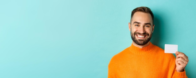 Primer plano de un apuesto hombre caucásico yendo de compras mostrando tarjeta de crédito y sonriendo de pie sobre tu