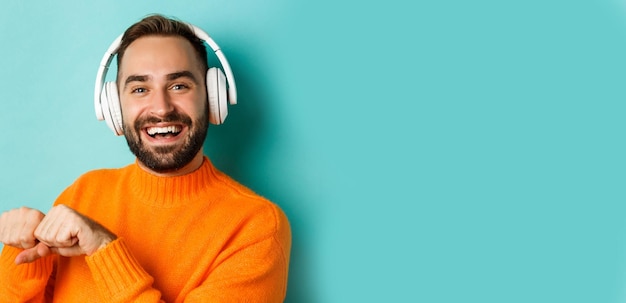 Foto gratuita primer plano de un apuesto hombre moderno escuchando música en auriculares de pie en un suéter naranja sobre turquesa