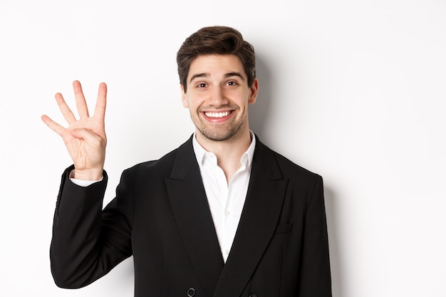 Primer plano de un apuesto hombre de negocios en traje negro, sonriendo asombrado, mostrando el número cuatro, de pie sobre fondo blanco.