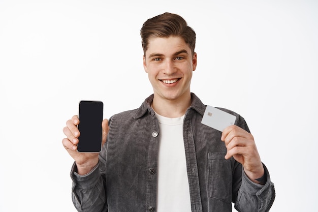 Primer plano de un apuesto hombre sonriente que muestra la tarjeta de crédito y la pantalla del teléfono inteligente, la interfaz de la aplicación de banca financiera, el pago en línea, las compras con la aplicación, el fondo blanco.