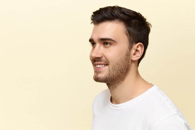 Primer plano de un apuesto joven caucásico confiado con cerdas y elegante corte de pelo posando aislado contra el fondo de pared de estudio de espacio de copia en blanco con alegre sonrisa feliz, de buen humor