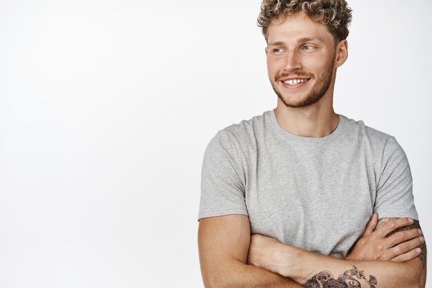 Primer plano de un apuesto joven rubio con tatuajes de cerdas y una sonrisa feliz cruzando los brazos en el pecho de pie en una pose relajada con una camiseta gris de fondo blanco